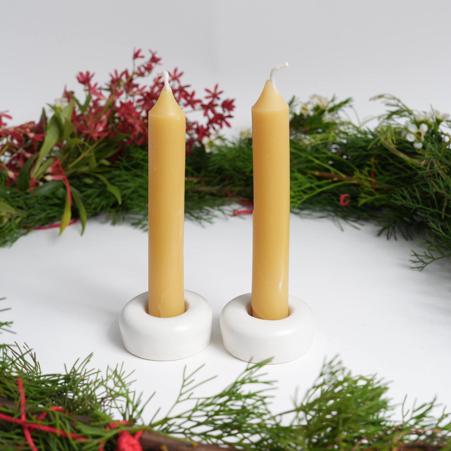 Two taper beeswax candles in white holders with a Christmas wreath in the background