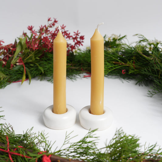 Two taper beeswax candles in white holders with a Christmas wreath in the background
